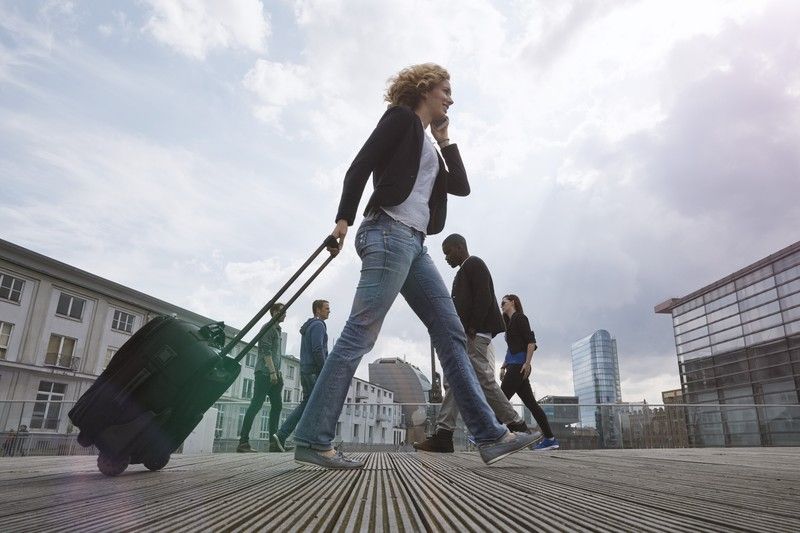 travail en mouvement Femme active en déplacement professionnel, tirant une valise à roulettes en centre urbain, symbole de la mobilité et des mutations du travail moderne.